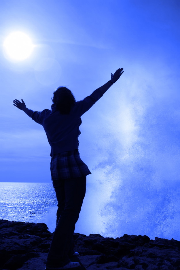 a lone woman raising her arms in awe at the powerful wave on the cliffs edge